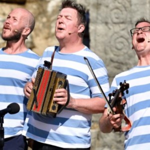 A photo of three men singing, all of them are wearing blue and white stripe tops. One is playing an accordion.