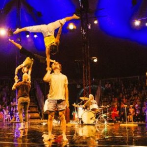 A photograph of a group of acrobats. One man holds a woman on his hand, she is upside down balancing on one hand. 