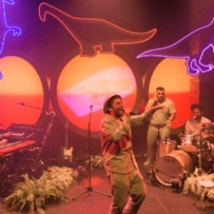 A photo of a man in a safari outfit singing into a microphone. In the background one man plays the drums and one man is standing. There are neon signs in the shape of dinosaurs in the background. 